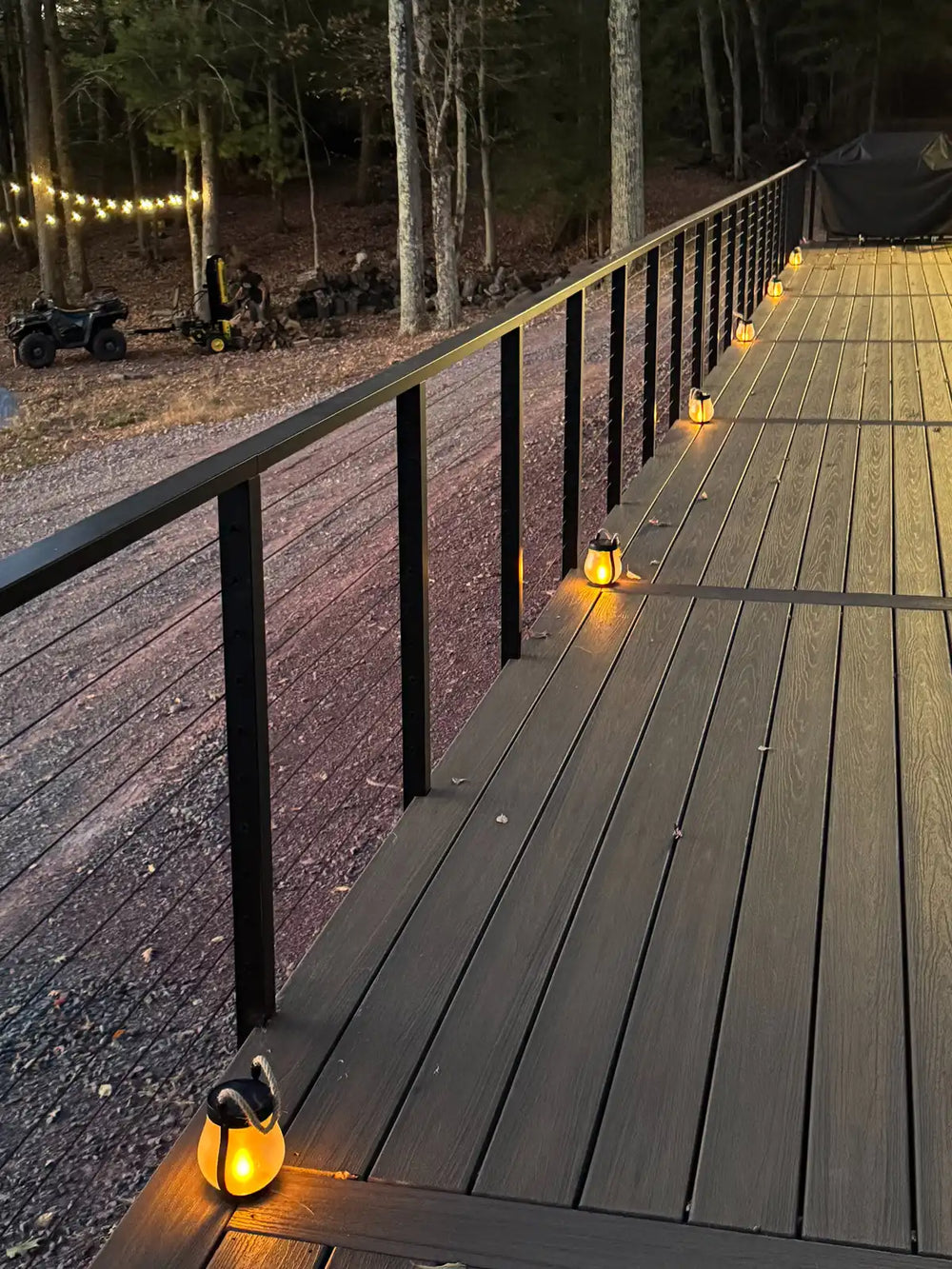 black deck railing illuminated by warm lantern lights in a wooded backyard.